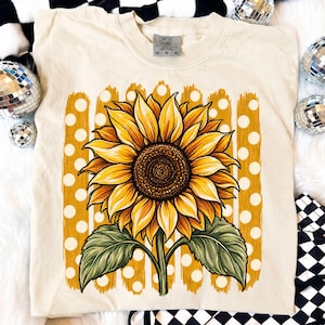 Könnte beinhalten: Ein cremefarbenes T-Shirt mit einer großen, detaillierten Sonnenblumengrafik mit gelben Blütenblättern und einer dunkelbraunen Mitte. Die Sonnenblume ist vor einem Hintergrund aus vertikalen gelben Pinselstrichen und weißen Punkten platziert.