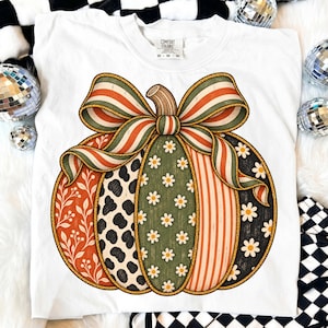 Puede incluir: Una camiseta blanca con un gráfico grande de una calabaza adornada con un lazo a rayas. La calabaza está dividida en secciones verticales con patrones que incluyen hojas naranjas, manchas animales en blanco y negro, margaritas y rayas.