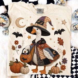 Può includere: Una t-shirt color crema con la grafica di un'oca che indossa un cappello da strega e un mantello patchwork. L'oca tiene in mano un secchiello a forma di zucca. Attorno all'oca ci sono illustrazioni di pipistrelli, stelle, luna e fiori.
