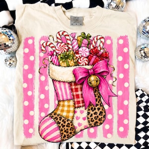 May include: A cream-colored t-shirt featuring a graphic of a Christmas stocking filled with candy canes, wrapped gifts, and peppermint candies. The stocking has a patchwork design with pink stripes, red plaid, yellow gingham, and leopard print.
