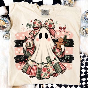 Op de afbeelding: Een crèmekleurig t-shirt met een afbeelding van een geest met een grote geruite strik, die een mok met een kerstboomontwerp en een peperkoekmannetje vasthoudt. De geest is versierd met een lichtsnoer en feestelijke patchworkpatronen.