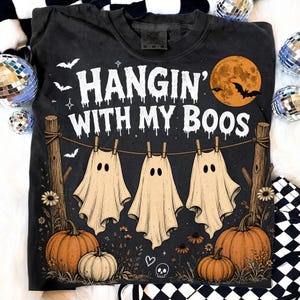 May include: A charcoal gray t-shirt featuring a graphic print with the text "HANGIN' WITH MY BOOS" in white, dripping letters. The design includes three white ghost sheets hanging on a clothesline between two wooden posts, pumpkins, and a moon.
