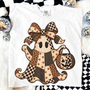 Op de afbeelding: Een wit t-shirt met een afbeelding van een geest met een grote strik. De geest heeft patronen van bruine luipaardprint, beige stippen en zwart-witte schaakbordpatronen. Het houdt een bijpassende pompoenvormige emmer voor snoep vast.