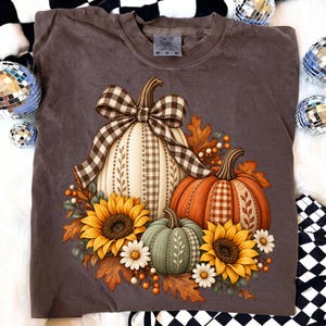 Puede incluir: Una camiseta marrón oscuro con un gráfico de tres calabazas en blanco, naranja y verde, decoradas con girasoles, margaritas blancas y hojas de otoño. Un gran lazo de cuadros marrón y blanco adorna la calabaza blanca central.
