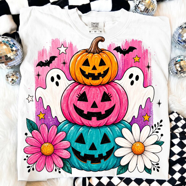 May include: White Comfort Colors t-shirt featuring a stacked pumpkin design with jack-o'-lantern faces in orange, pink, and teal. The design includes ghosts, bats, stars, and flowers, perfect for Halloween.