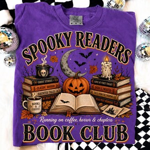 May include: A purple t-shirt featuring a graphic of a jack-o'-lantern, an open book, stacks of books, a candle, and mugs. The text reads SPOOKY READERS, BOOK CLUB, and Running on coffee, horror & chapters. The design includes bats, leaves, and a crescent moon.