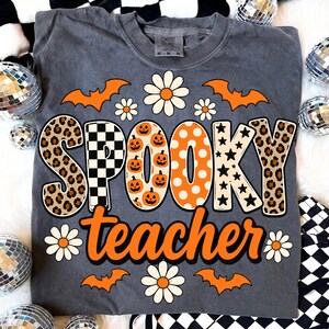 K&ouml;nnte beinhalten: Ein anthrazitfarbenes T-Shirt mit dem Schriftzug SPOOKY teacher in gro&szlig;en, gemusterten Buchstaben. Das Design zeigt orangefarbene K&uuml;rbisse, schwarz-wei&szlig;e Karos, Leopardenmuster und Sterne, erg&auml;nzt durch orangefarbene Flederm&auml;use und wei&szlig;e G&auml;nsebl&uuml;mchen.