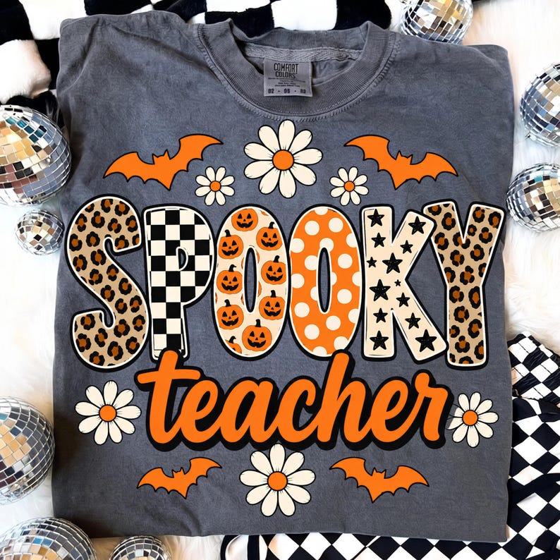 K&ouml;nnte beinhalten: Ein anthrazitfarbenes T-Shirt mit dem Schriftzug SPOOKY teacher in gro&szlig;en, gemusterten Buchstaben. Das Design zeigt orangefarbene K&uuml;rbisse, schwarz-wei&szlig;e Karos, Leopardenmuster und Sterne, erg&auml;nzt durch orangefarbene Flederm&auml;use und wei&szlig;e G&auml;nsebl&uuml;mchen.