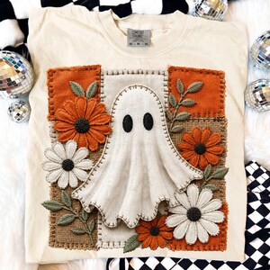 May include: A cream-colored t-shirt featuring a central rectangular patch with a ghost appliqué, surrounded by embroidered orange and white daisy-like flowers and green leaves on a textured burlap-style background with visible stitching.