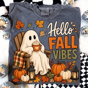 Puede incluir: Una camiseta gris carbón con un gráfico de un fantasma blanco sentado en una silla de madera, sosteniendo una taza con un corazón. El texto dice Hello FALL VIBES. El diseño incluye calabazas naranjas, hojas de otoño y dos faroles negros.