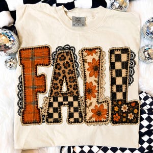 May include: A cream-colored t-shirt featuring the word FALL in large block letters. Each letter has a unique pattern: orange plaid, leopard print, orange flowers, and black and white checkers. The letters are outlined with black lace trim.