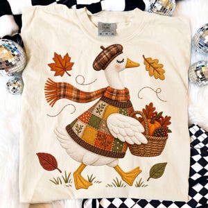 Puede incluir: Una camiseta color crema con el gráfico de un ganso blanco que lleva una boina y bufanda de cuadros marrones. El ganso viste un suéter de retazos y lleva una cesta tejida llena de una calabaza, una piña y hojas, rodeado de hojas de otoño.