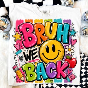 May include: A white t-shirt featuring a colorful graphic with the text BRUH WE BACK. The design includes a yellow smiley face, a yellow pencil, a red apple, a pink eraser, a notebook page, stars, hearts, and a flower over vertical rainbow stripes.