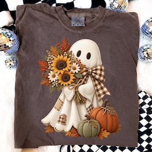 Può includere: Una t-shirt marrone a maniche corte con la grafica di un fantasma bianco che tiene in mano un bouquet di girasoli, margherite e foglie autunnali. Il fantasma indossa un fiocco a quadri marrone e bianco accanto a una zucca verde e arancione.