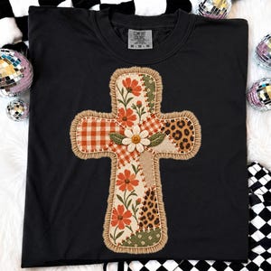 May include: A black t-shirt featuring a large, central cross-shaped applique. The cross is composed of various fabric patches including orange floral, orange gingham, leopard print, and green patterns, all outlined with a thick, frayed tan fringe.