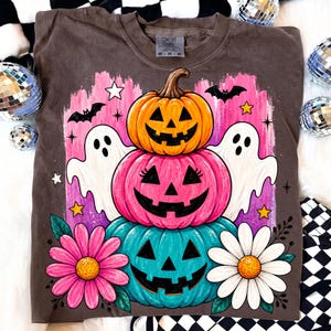 May include: A brown Comfort Colors sweatshirt featuring a stacked pumpkin design with jack-o'-lantern faces in orange, pink, and teal. The design includes white ghosts, bats, stars, and flowers, perfect for Halloween.