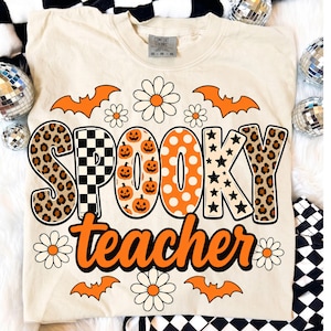 K&ouml;nnte beinhalten: Ein cremefarbenes T-Shirt mit dem Schriftzug SPOOKY in gro&szlig;en, gemusterten Buchstaben, darunter Leopardenmuster, schwarz-wei&szlig;es Karomuster, orangefarbene K&uuml;rbisse, wei&szlig;e Punkte und schwarze Sterne, mit dem Wort teacher in Orange.