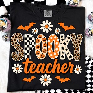 K&ouml;nnte beinhalten: Ein schwarzes T-Shirt mit dem Text SPOOKY in gro&szlig;en, gemusterten Buchstaben, darunter Leopardenmuster, schwarz-wei&szlig;e Karos, orangefarbene K&uuml;rbisse, orangefarbene Punkte und schwarze Sterne. Das Wort teacher steht darunter in Orange.