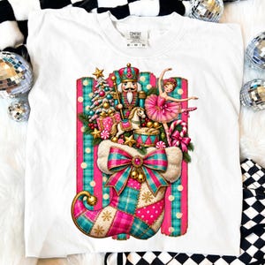 May include: A white t-shirt featuring a graphic of a Christmas stocking filled with a nutcracker, a ballerina, a decorated tree, a rocking horse, and a drum. The stocking is patterned with pink and teal plaid and polka dots, finished with a large bow.
