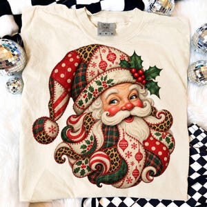 May include: A cream-colored t-shirt featuring a graphic of Santa Claus with a long, curly beard and a hat. The design incorporates patchwork patterns including red polka dots, green plaid, and leopard print, accented with holly leaves and berries.