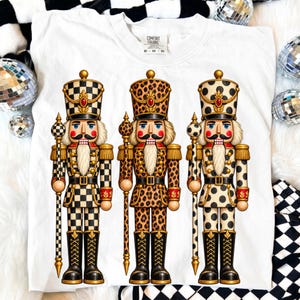 May include: A white t-shirt featuring a graphic print of three nutcracker figures. The first nutcracker has a black and white checkered pattern, the second has a leopard print, and the third has a black polka dot pattern on a cream background.