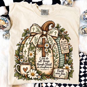 Puede incluir: Una camiseta de color crema con un gráfico de una calabaza decorada con un lazo grande, una cruz de madera, margaritas y vegetación. El texto dice: I can do all things through Christ who strengthens me, Be still and know that I am God, y Faith over fear.