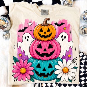May include: Cream-colored sweatshirt featuring a stacked pumpkin design with jack-o'-lantern faces in orange, pink, and teal. Includes white ghosts, bats, stars, and flowers. The shirt has the text "Comfort Colors" on the tag.