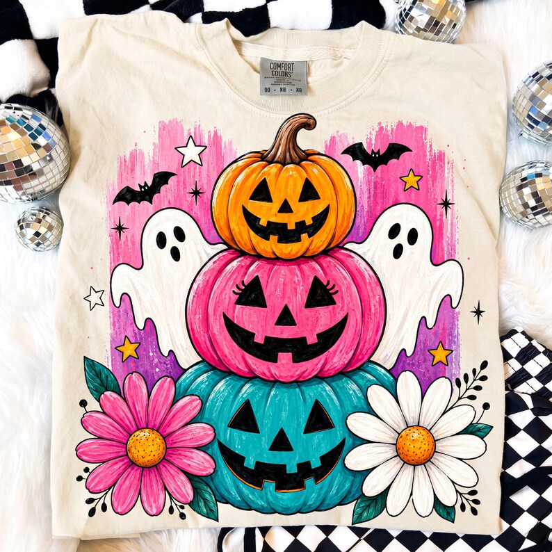 May include: Cream-colored sweatshirt featuring a stacked pumpkin design with jack-o'-lantern faces in orange, pink, and teal. Includes white ghosts, bats, stars, and flowers. The shirt has the text "Comfort Colors" on the tag.