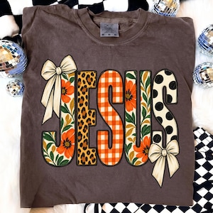 May include: A brown short-sleeve t-shirt featuring the word JESUS in large letters. Each letter has a unique pattern: floral with orange flowers, leopard print, orange and white gingham, floral, and white with black polka dots, plus two bows.