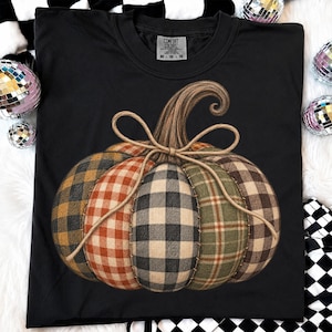 May include: A black short-sleeved t-shirt featuring a large graphic of a pumpkin made from various plaid fabric segments in shades of yellow, orange, black, and green. The pumpkin is tied with a tan twine bow and has a brown stem.