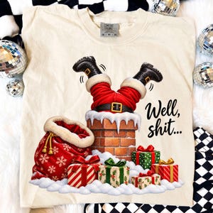 May include: A cream-colored t-shirt featuring a graphic of Santa Claus upside down in a brick chimney. The design includes a red gift sack, wrapped presents, snow, and the black text "Well, shit..." printed on the right side of the shirt.