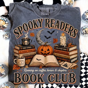 Op de afbeelding: Een antracietgrijs t-shirt met een grafische print met de tekst "SPOOKY READERS" en "BOOK CLUB". Het ontwerp bevat stapels boeken, een pompoen, een maansikkel, vleermuizen, een kaars en mokken met de tekst "BOOKS & COFFEE".