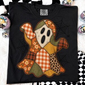 May include: A black short-sleeved t-shirt featuring a large ghost graphic made of various patchwork patterns including orange gingham, floral prints, leopard spots, and solid olive green. The design includes black spiderweb illustrations.