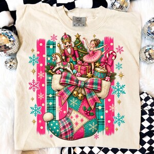 Puede incluir: Una camiseta de color crema con un gráfico de un calcetín navideño lleno de un cascanueces, una bailarina, un caballito de madera, una caja de regalo y un bastón de caramelo. El diseño incluye patrones de cuadros rosas y turquesas.