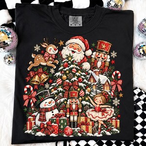 Puede incluir: Una camiseta negra con un gráfico navideño festivo que muestra a Santa Claus, un reno, un muñeco de nieve, dos cascanueces y una bailarina. El diseño incluye un árbol decorado, regalos envueltos, bastones de caramelo y copos.