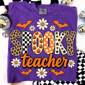 K&ouml;nnte beinhalten: Ein violettes Kurzarm-T-Shirt mit dem Schriftzug SPOOKY teacher in gro&szlig;en, gemusterten Buchstaben. Das Wort SPOOKY enth&auml;lt Leopardenmuster, schwarz-wei&szlig;e Karos, K&uuml;rbisse, orangefarbene Punkte und schwarze Sterne. Orangefarbene Flederm&auml;use und wei&szlig;e G&auml;nsebl&uuml;mchen umgeben den Text.
