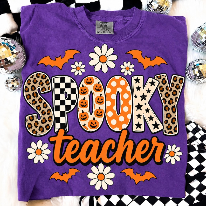 K&ouml;nnte beinhalten: Ein violettes Kurzarm-T-Shirt mit dem Schriftzug SPOOKY teacher in gro&szlig;en, gemusterten Buchstaben. Das Wort SPOOKY enth&auml;lt Leopardenmuster, schwarz-wei&szlig;e Karos, K&uuml;rbisse, orangefarbene Punkte und schwarze Sterne. Orangefarbene Flederm&auml;use und wei&szlig;e G&auml;nsebl&uuml;mchen umgeben den Text.