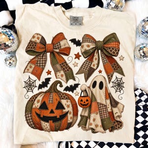 Puede incluir: Una camiseta color crema con un estampado gráfico de dos grandes lazos estilo patchwork, una calabaza de Halloween y un fantasma que sostiene un pequeño cubo de calabaza. El diseño incluye pequeñas estrellas, telarañas y murciélagos.