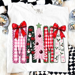 May include: A white t-shirt featuring the word MAMA in large, decorative letters. The letters are decorated with red and pink gingham patterns, a green background with snowmen, a pink Christmas tree, and holly leaves with red bows.