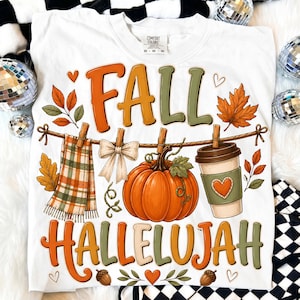 Puede incluir: Una camiseta blanca con un estampado gráfico que incluye el texto FALL HALLELUJAH en naranja y verde. El diseño muestra una bufanda de cuadros, un lazo crema, una calabaza y una taza de café colgados de un tendedero, con hojas de otoño y corazones.