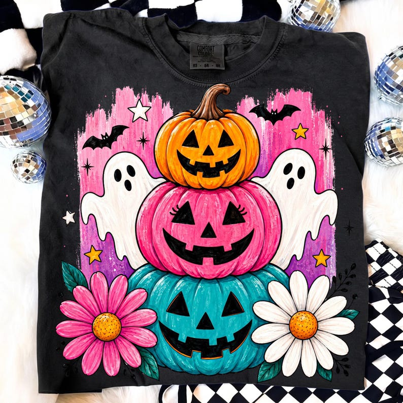May include: Black crew neck t-shirt featuring a stacked pumpkin design with a jack-o'-lantern face. The design includes pink, orange, and teal pumpkins, white ghosts, bats, stars, and flowers. The shirt is a Comfort Colors brand.