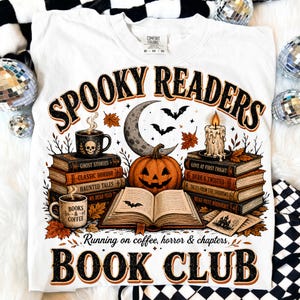 May include: A white t-shirt featuring a graphic print with the text "SPOOKY READERS" and "BOOK CLUB". The design includes stacks of books, a jack-o'-lantern, a crescent moon, bats, a lit candle, and two mugs with the text "BOOKS & COFFEE".