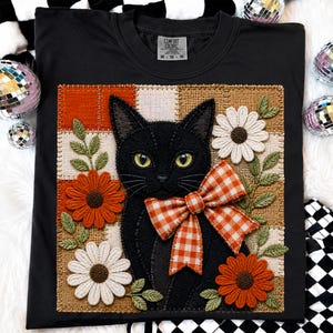 Könnte beinhalten: Ein schwarzes T-Shirt mit einem quadratischen Aufnäher, der eine gestickte schwarze Katze mit einer orange-weiß karierten Schleife zeigt. Der Aufnäher enthält orange und weiße Blumen, grüne Blätter und einen karierten Hintergrund in Orange- und Beigetönen.