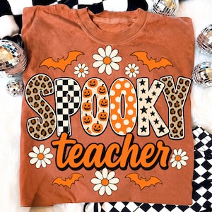K&ouml;nnte beinhalten: Ein rostfarbenes T-Shirt mit dem Schriftzug SPOOKY teacher in gro&szlig;en, gemusterten Buchstaben. Das Design zeigt orangefarbene K&uuml;rbisse, schwarz-wei&szlig;e Karos, Leopardenmuster, Sterne und Punkte, umgeben von wei&szlig;en G&auml;nsebl&uuml;mchen und orangefarbenen Flederm&auml;usen.
