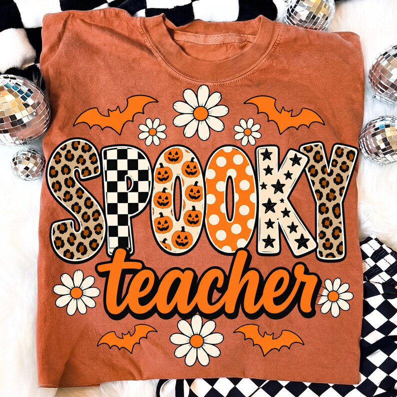 K&ouml;nnte beinhalten: Ein rostfarbenes T-Shirt mit dem Schriftzug SPOOKY teacher in gro&szlig;en, gemusterten Buchstaben. Das Design zeigt orangefarbene K&uuml;rbisse, schwarz-wei&szlig;e Karos, Leopardenmuster, Sterne und Punkte, umgeben von wei&szlig;en G&auml;nsebl&uuml;mchen und orangefarbenen Flederm&auml;usen.