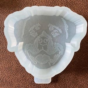 May include: A light blue, dog-shaped mold with a detailed face design. The mold features a raised border and a small tab at the bottom. The dog's face is etched into the mold, showcasing its eyes, nose, and mouth.