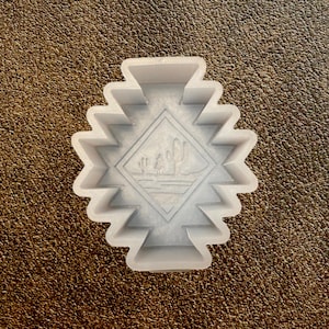 May include: A white plastic cookie cutter in a geometric design with a diamond-shaped center featuring a desert landscape with cacti. The cutter has a zig-zag edge, perfect for creating themed cookies.