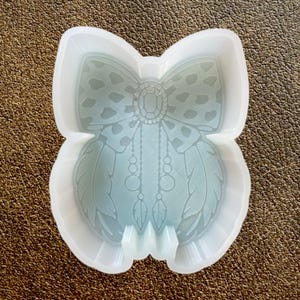 May include: A clear, flexible silicone mold in the shape of a dreamcatcher. The mold features a bow at the top, a central jewel, beaded strands, and feather details. The mold is designed for crafting and resin art projects.