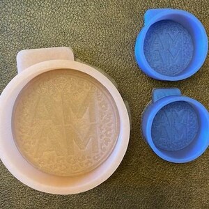 May include: Two blue silicone molds with the letters "AM" embossed in the center. One mold is larger and has a textured surface. The other mold is smaller and has a smooth surface.