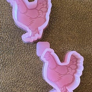 May include: Two pink silicone molds shaped like chickens. The molds are detailed with feathers and have a raised design.